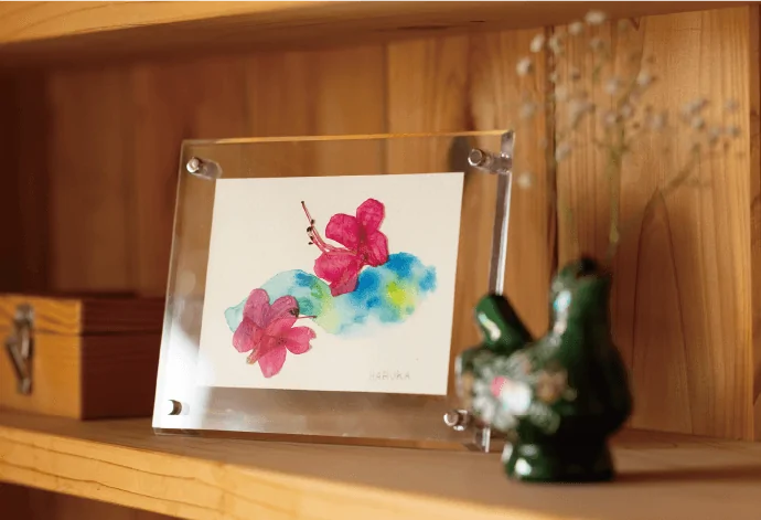 写真：木の飾り棚に置かれたピンク色の花の押し花アート作品と蛙の置物。